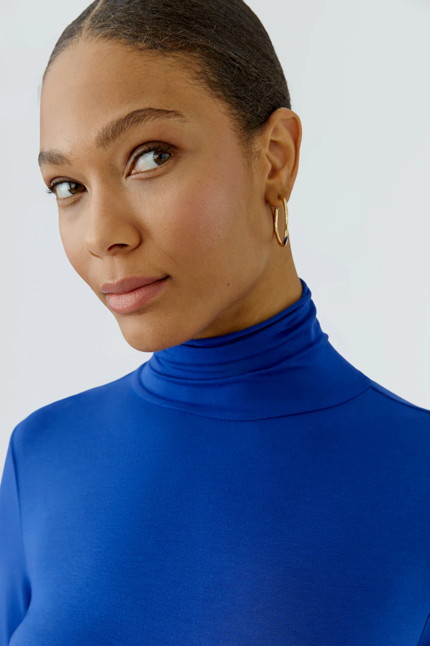 Woman in a blue long-sleeve shirt with a turtleneck, looking confidently to the side.