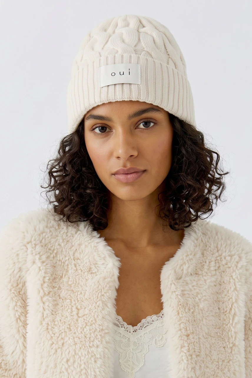 Woman in a white knit beanie, olive jacket, and casual jeans, showcasing a stylish winter look.