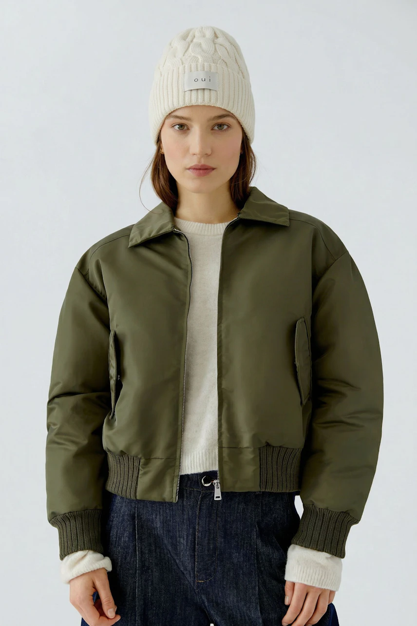 Frau mit cremefarbener Mütze von Oui, kombiniert mit olivgrüner Jacke und weißem Pullover.