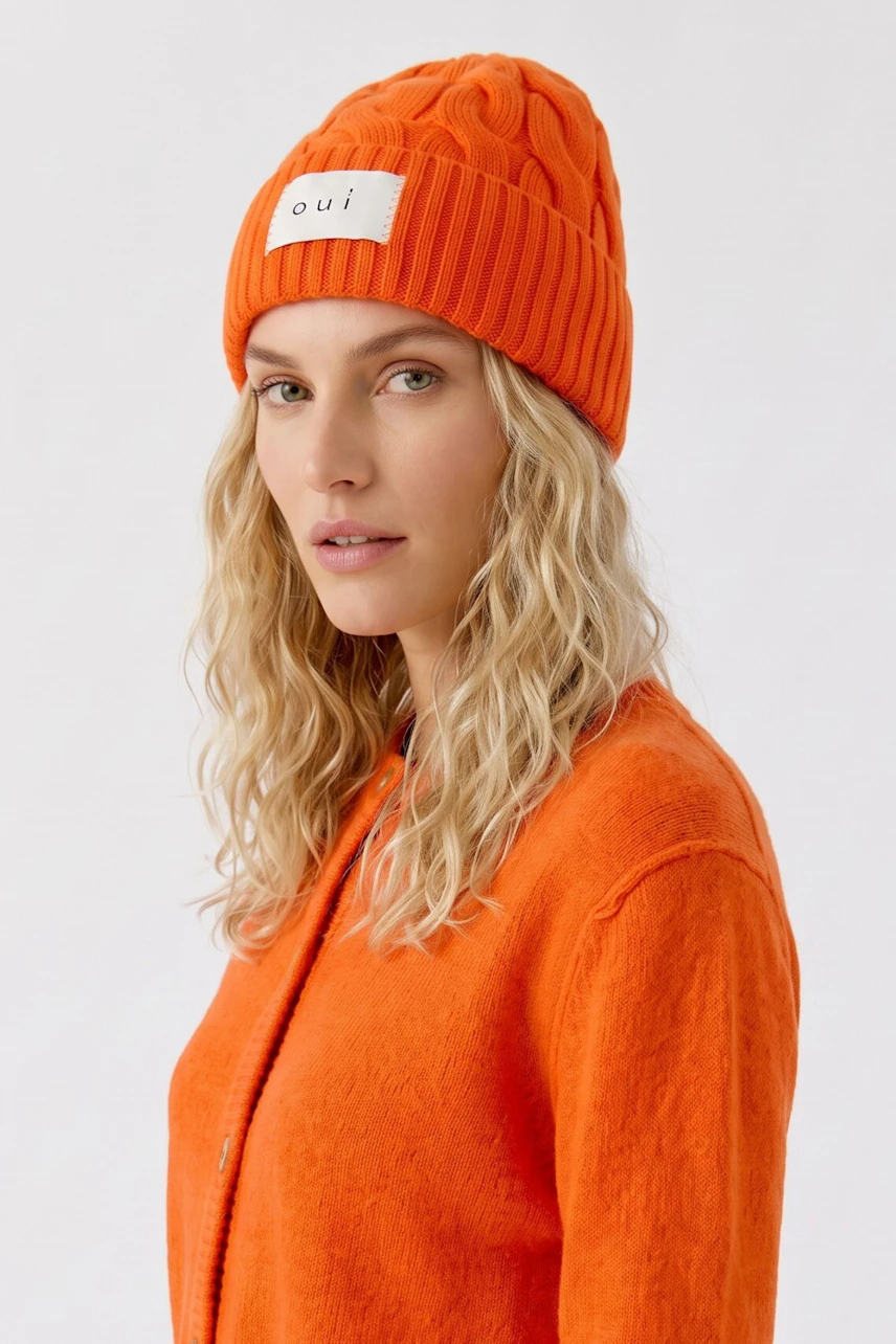 Frau mit orangefarbener Mütze und olivgrüner Jacke, seitlich blickend. Schlichtes, modernes Design.