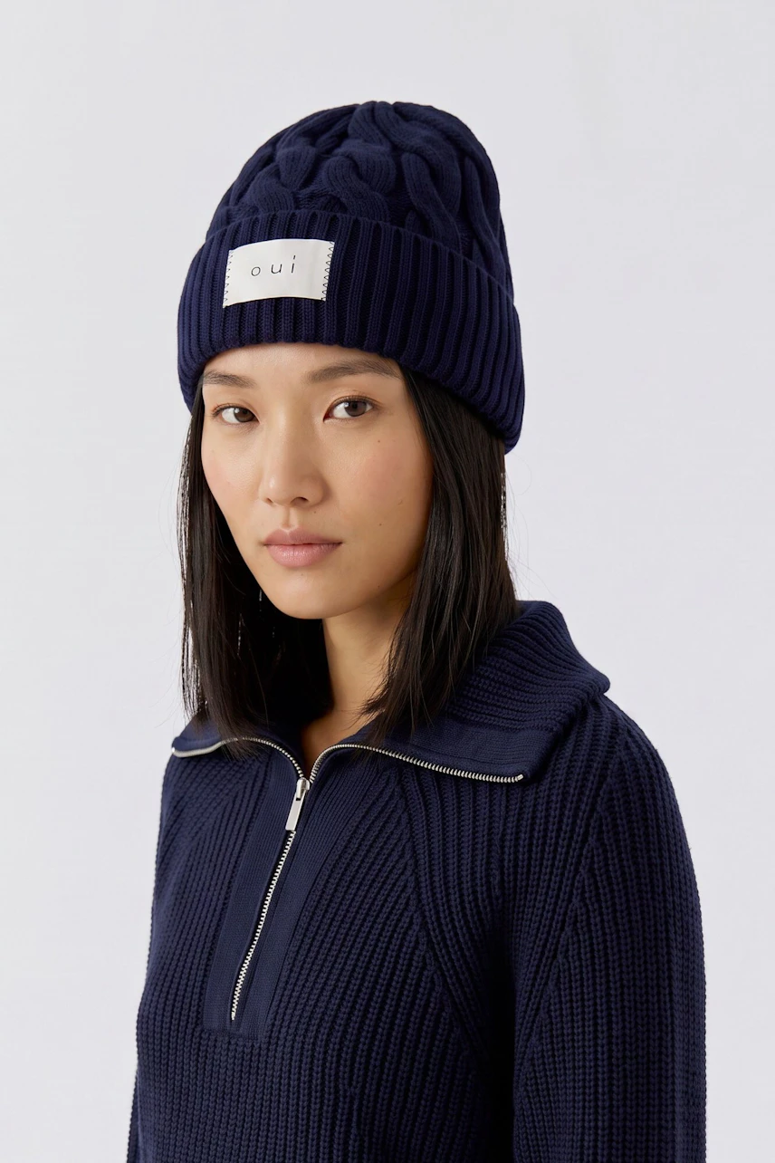 Woman in a navy knit pullover and matching beanie, looking confidently at the camera.