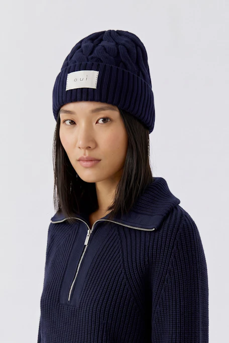 Dunkelblaue Strickmütze mit Zopfmuster und Oui-Logo, ideal für kalte Tage.