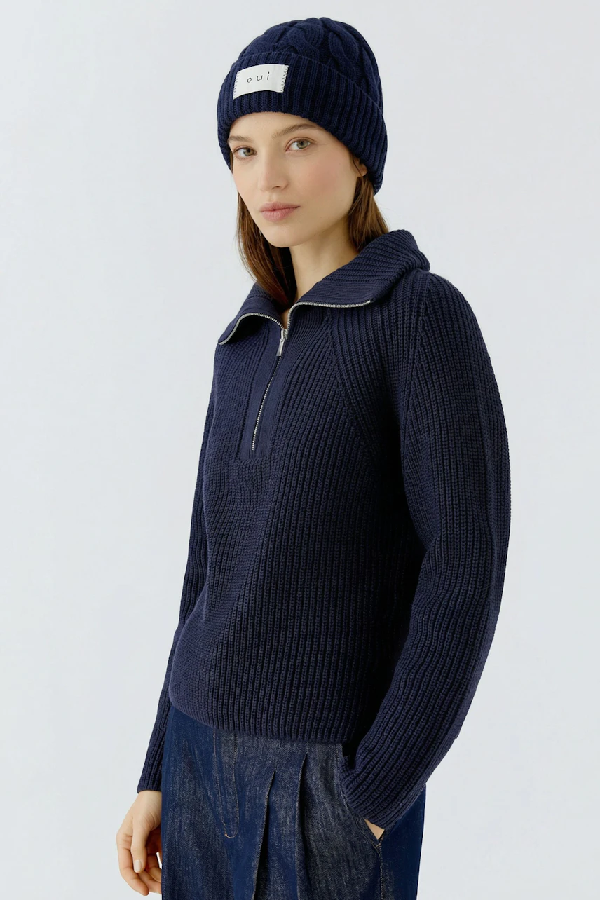 Damen-Mütze: Frau trägt dunkelblaue Strickmütze mit Logo, passend zum Pullover.