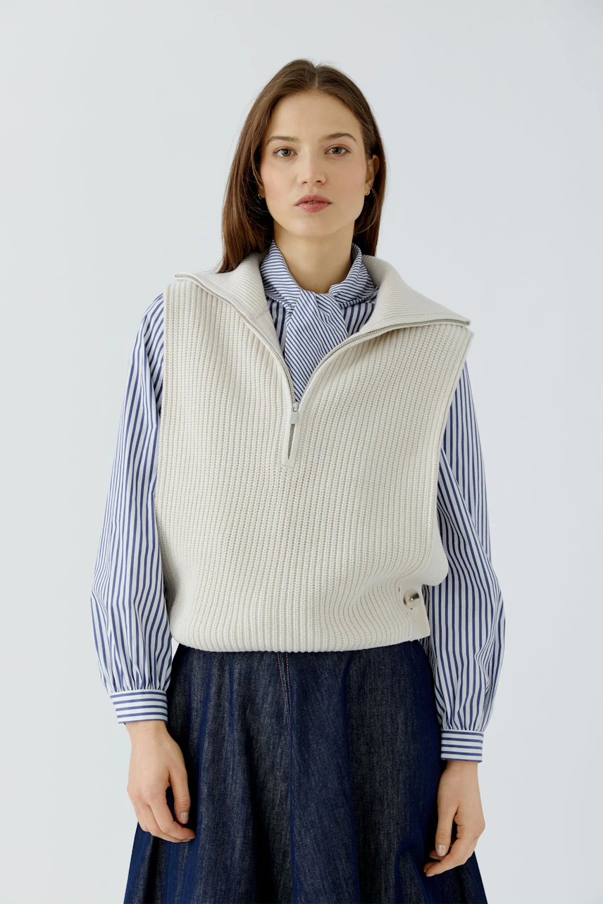 Casual women's pullover: Woman in a cream knit pullover over a striped shirt and denim skirt, looking poised.