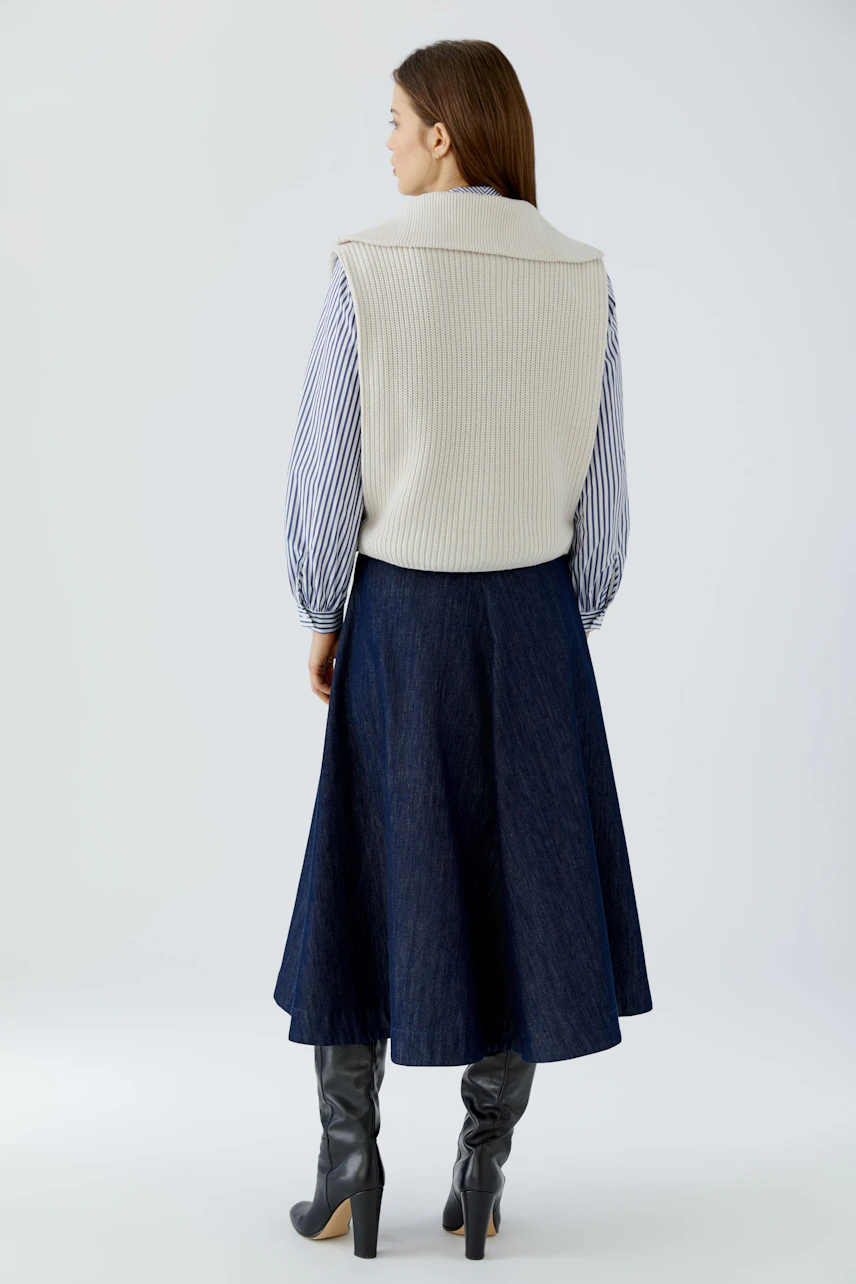Woman in a casual pullover vest over a striped shirt, paired with a denim skirt and knee-high boots.