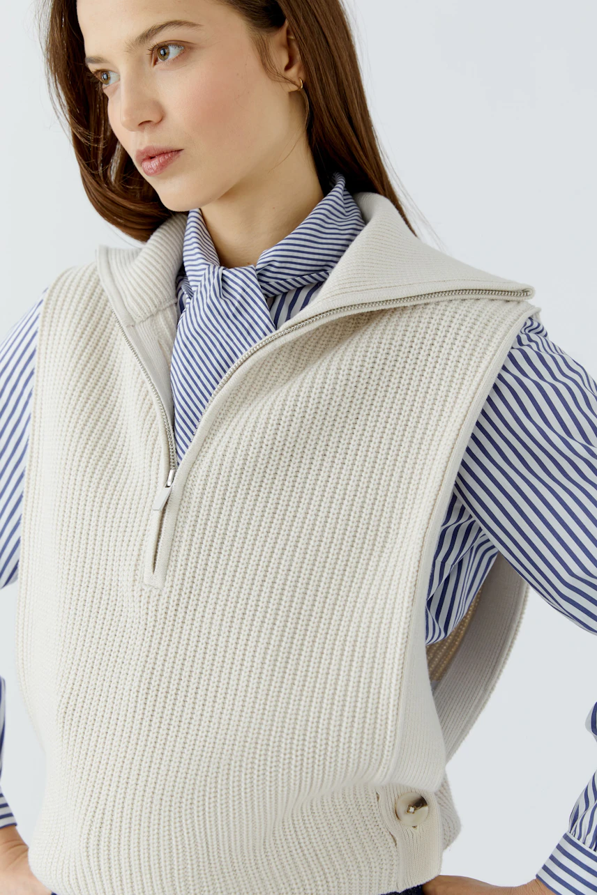 Casual women's pullover: Woman in a cream knit pullover over a blue striped shirt, looking to the side.