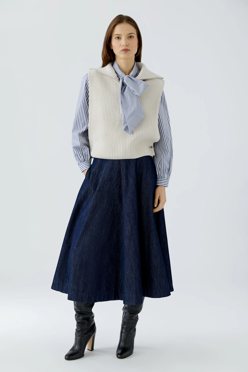 Woman in a striped shirt and white pullover vest, paired with a long denim skirt and black boots.