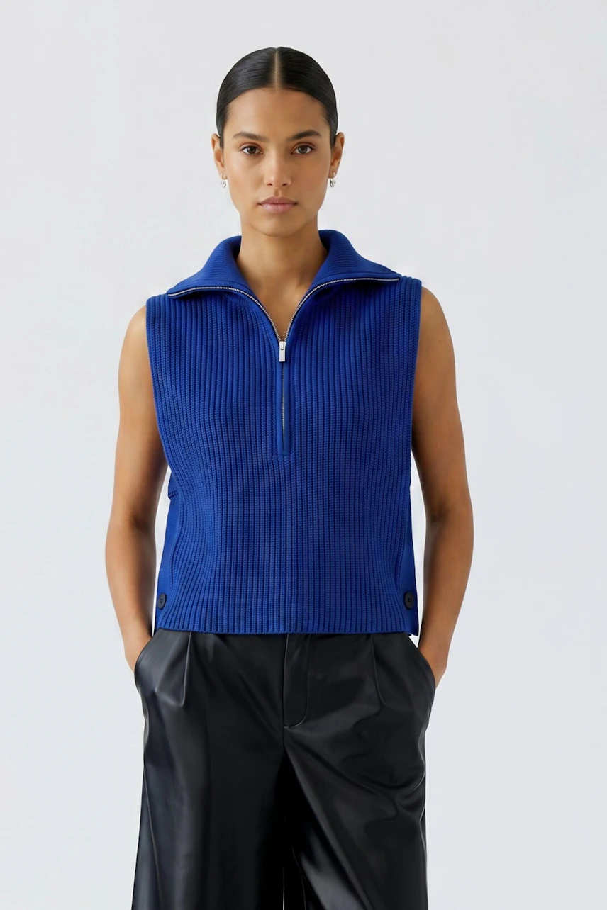 Woman in blue pullover vest with zip collar, paired with black pants, standing confidently.