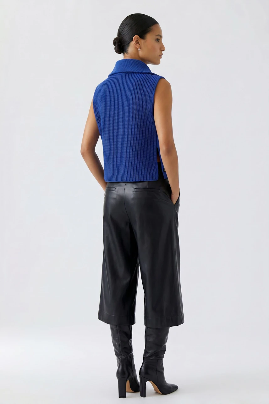 Woman in blue pullover vest, black leather culottes, and boots, standing confidently.