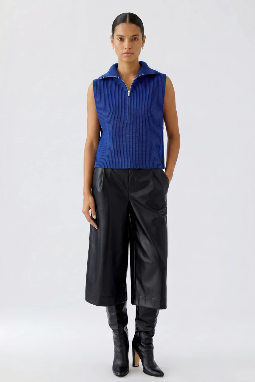 Woman in blue sleeveless pullover with zip, paired with black culottes and boots, standing confidently.
