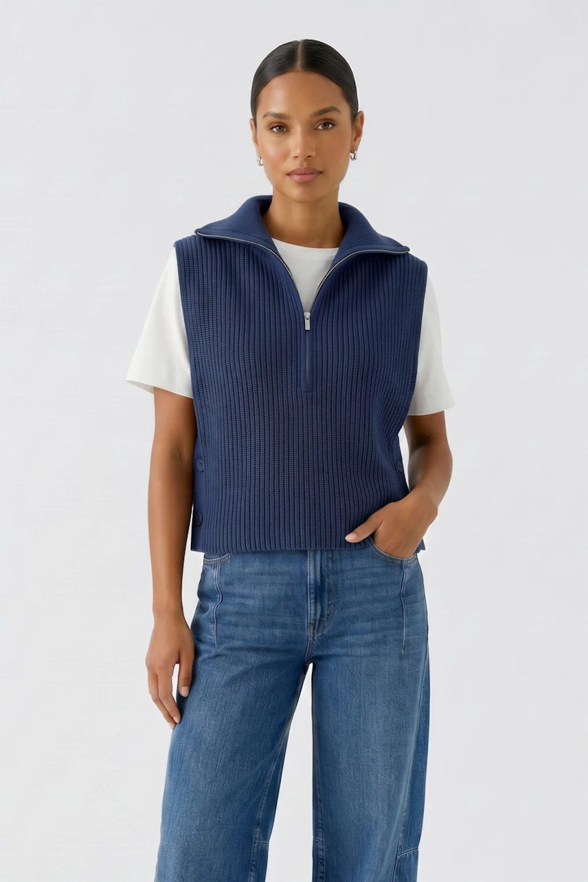 Woman in a blue pullover vest over a white shirt and jeans, standing confidently.