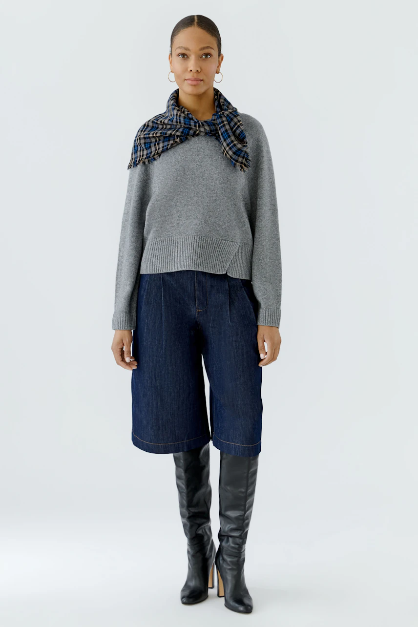 Frau mit grauem Pullover und kariertem Tuch, kombiniert mit Jeans-Shorts und schwarzen Stiefeln.