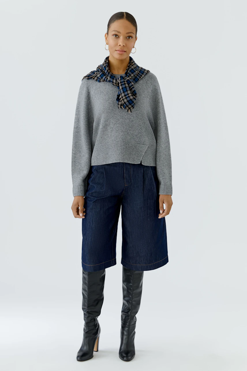 Frau mit kariertem Tuch um den Hals, grauem Pullover, Jeans-Culottes und schwarzen Stiefeln.