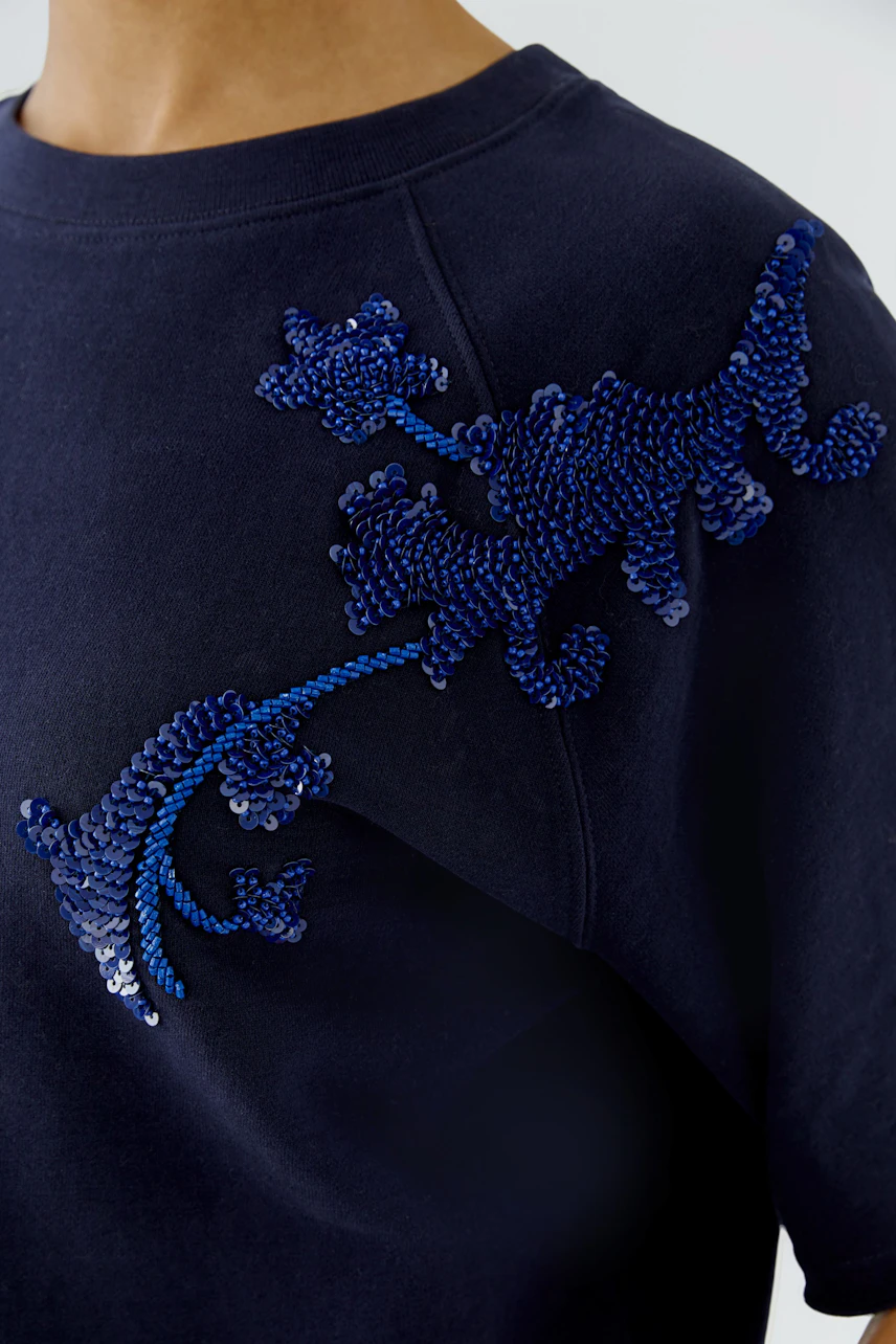 Navy sweatshirt with blue sequin floral design on shoulder, worn by a woman.