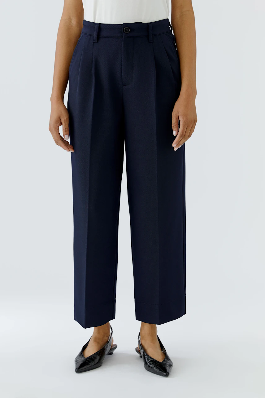 Woman in navy pleated trousers, white top, and black shoes, showcasing a relaxed, stylish look.