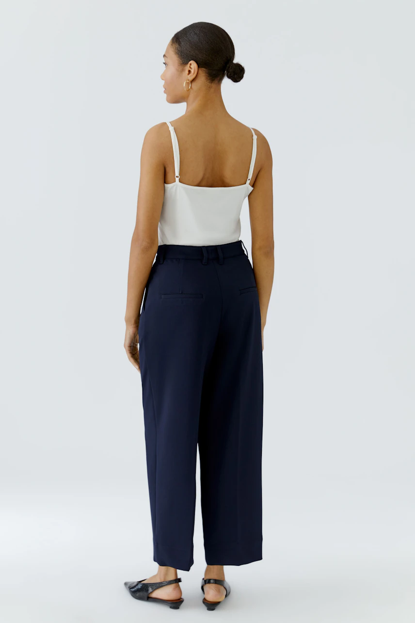 Woman in navy pleated trousers and white top, viewed from behind, showcasing relaxed fit and style.
