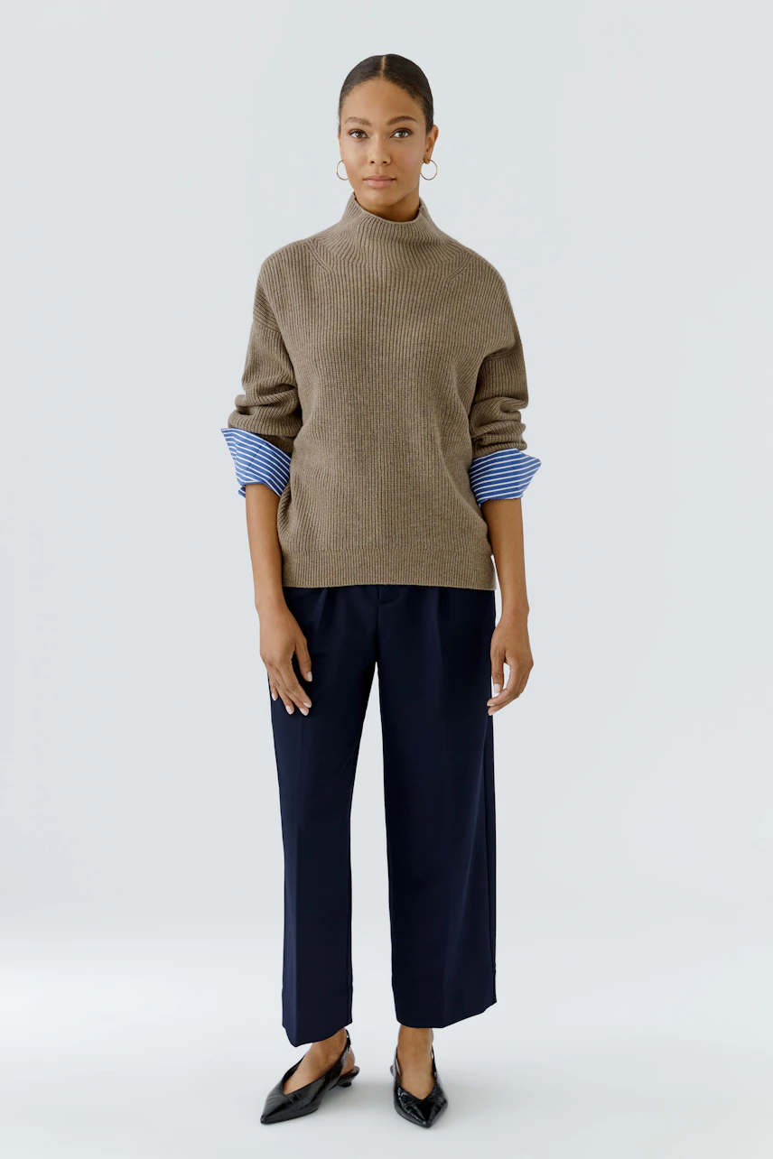 Woman in pleated trousers and a cozy brown pullover with blue striped cuffs, standing confidently.