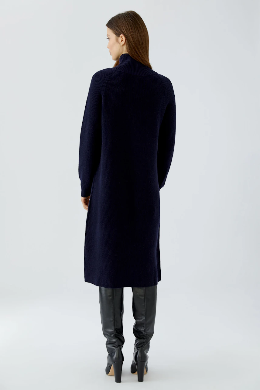 Woman in navy knit dress, long sleeves, standing confidently in black knee-high boots.