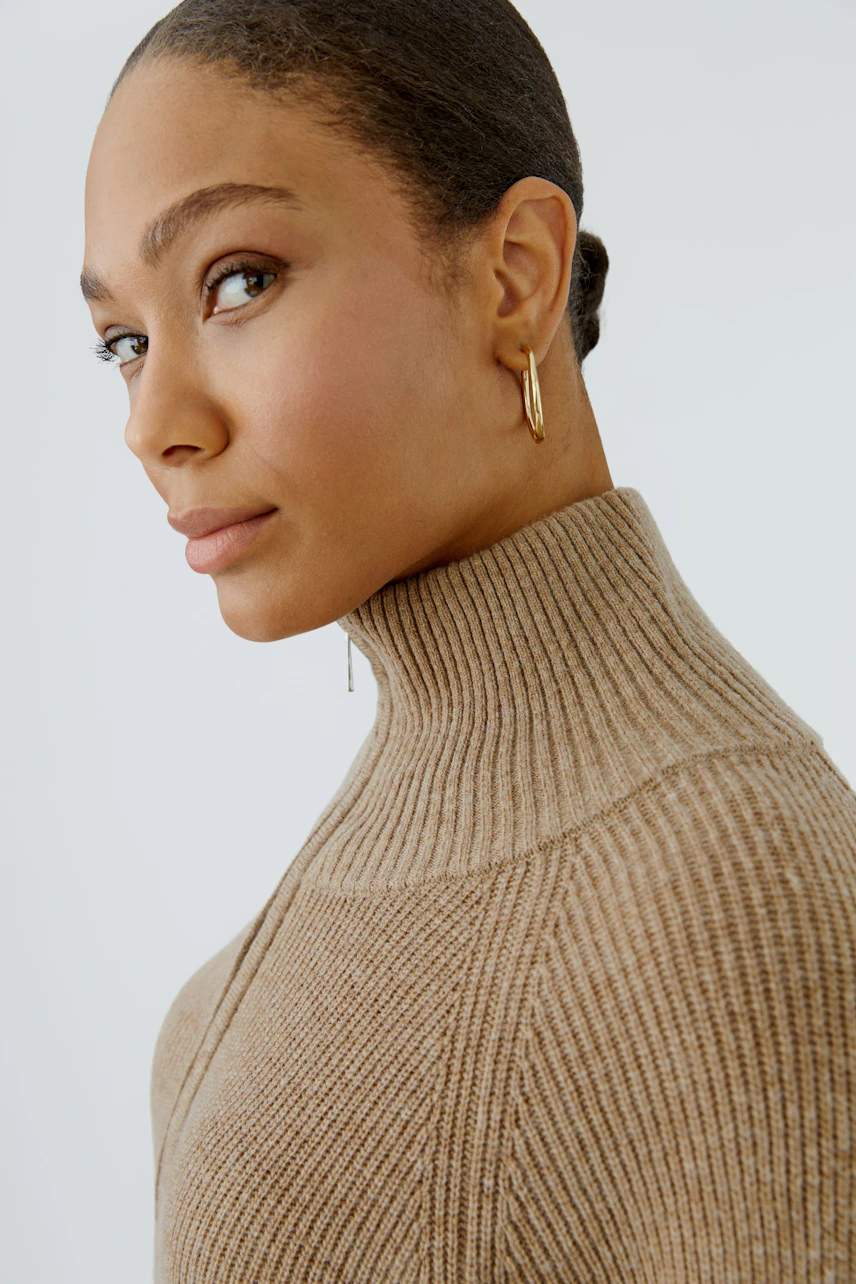 Woman in a beige knit dress with a high collar, looking confidently to the side.
