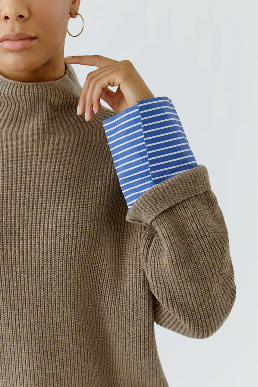 Woman in a brown ribbed pullover with blue striped cuffs, touching her face thoughtfully.