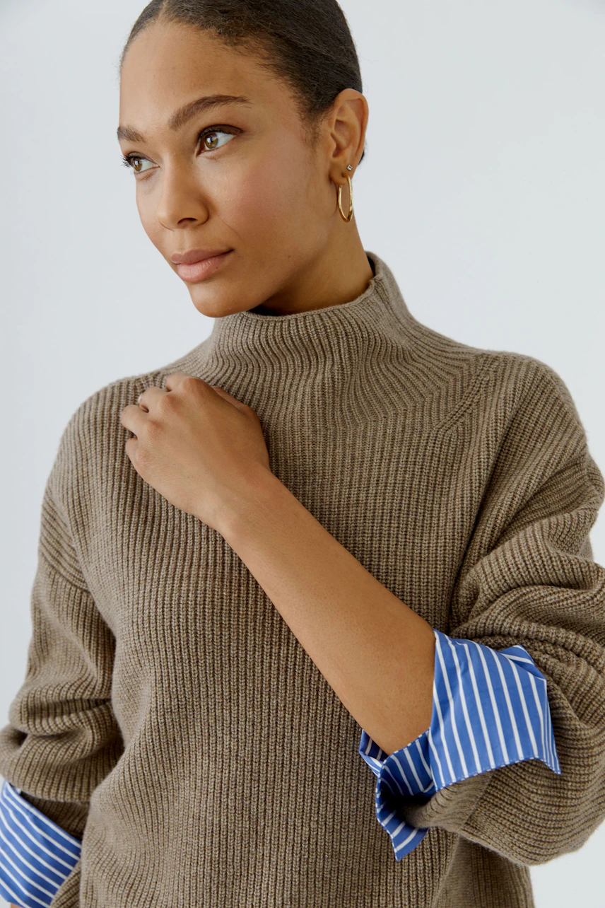 Casual women's pullover: Woman in a brown ribbed pullover with blue striped cuffs, looking thoughtful.
