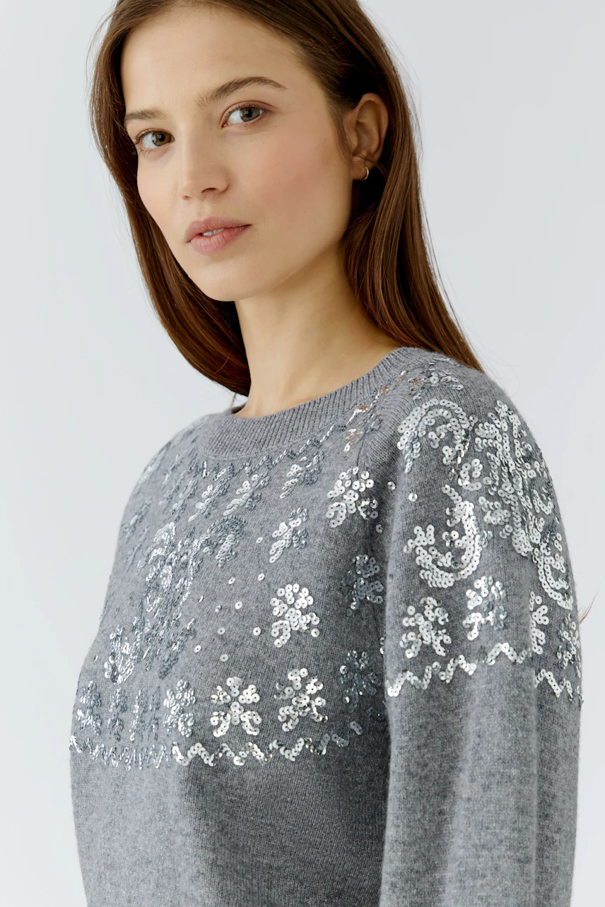 Woman in a grey pullover with intricate sequin patterns, looking confidently.