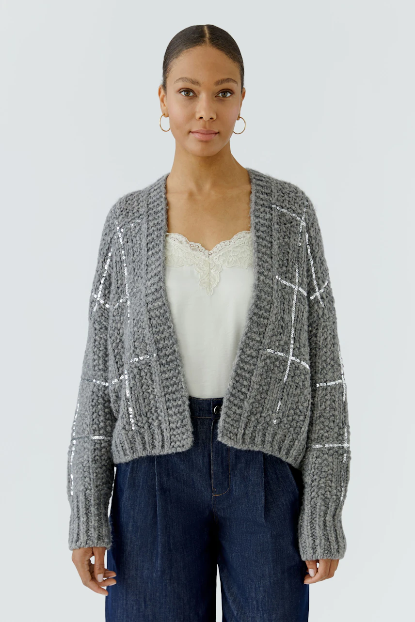 Casual knit cardigan: Woman in a grey chunky cardigan over a white top and blue jeans, looking relaxed.