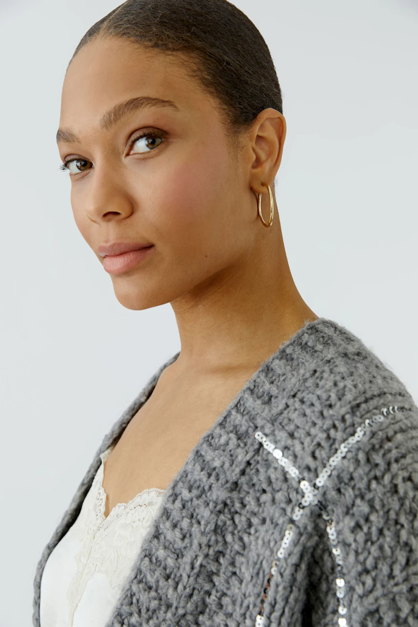 Woman in a cozy knit cardigan with sequins, wearing a white lace top, looking confidently to the side.