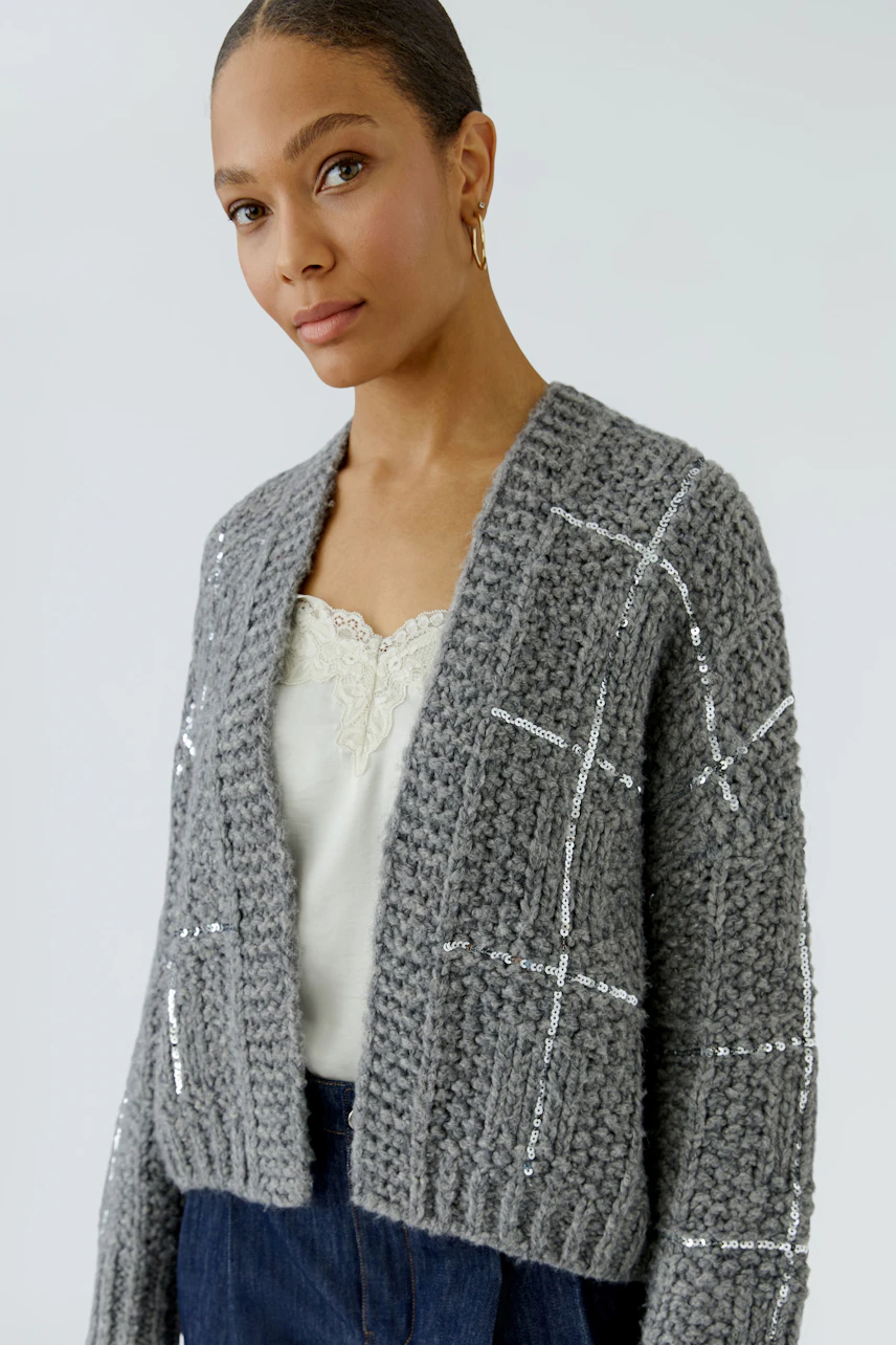 Woman in a grey knit cardigan with sequin details, wearing a white top and blue jeans.