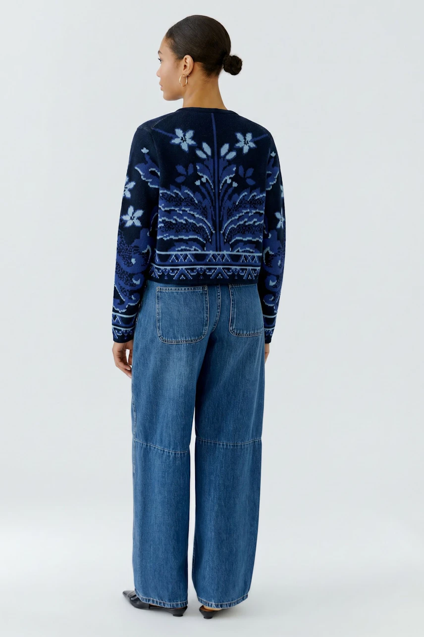 Frau in blauer Strickjacke mit floralem Muster, kombiniert mit lässiger Jeans, von hinten gesehen.