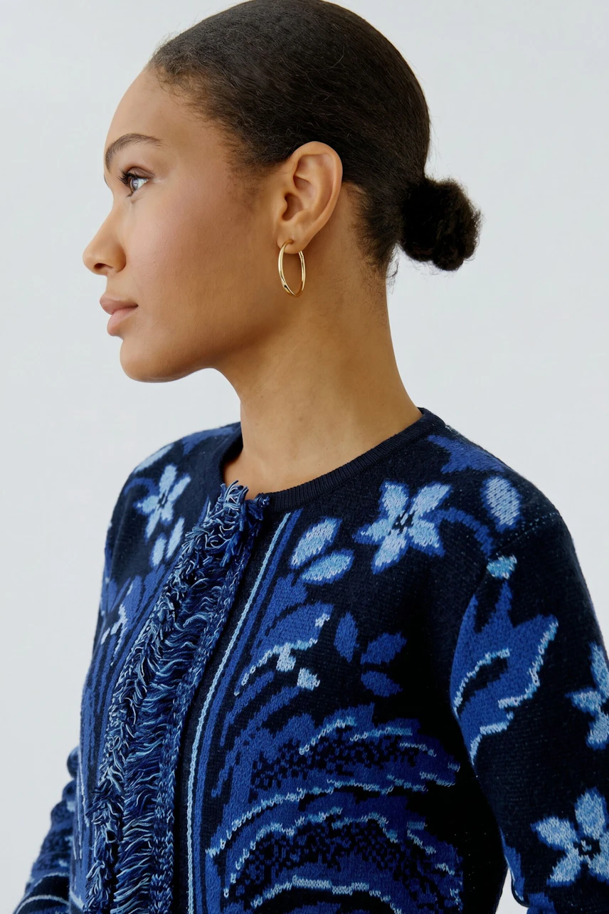 Strickjacke: Frau in blauer Strickjacke mit floralem Muster und goldenen Ohrringen, seitlich abgebildet.