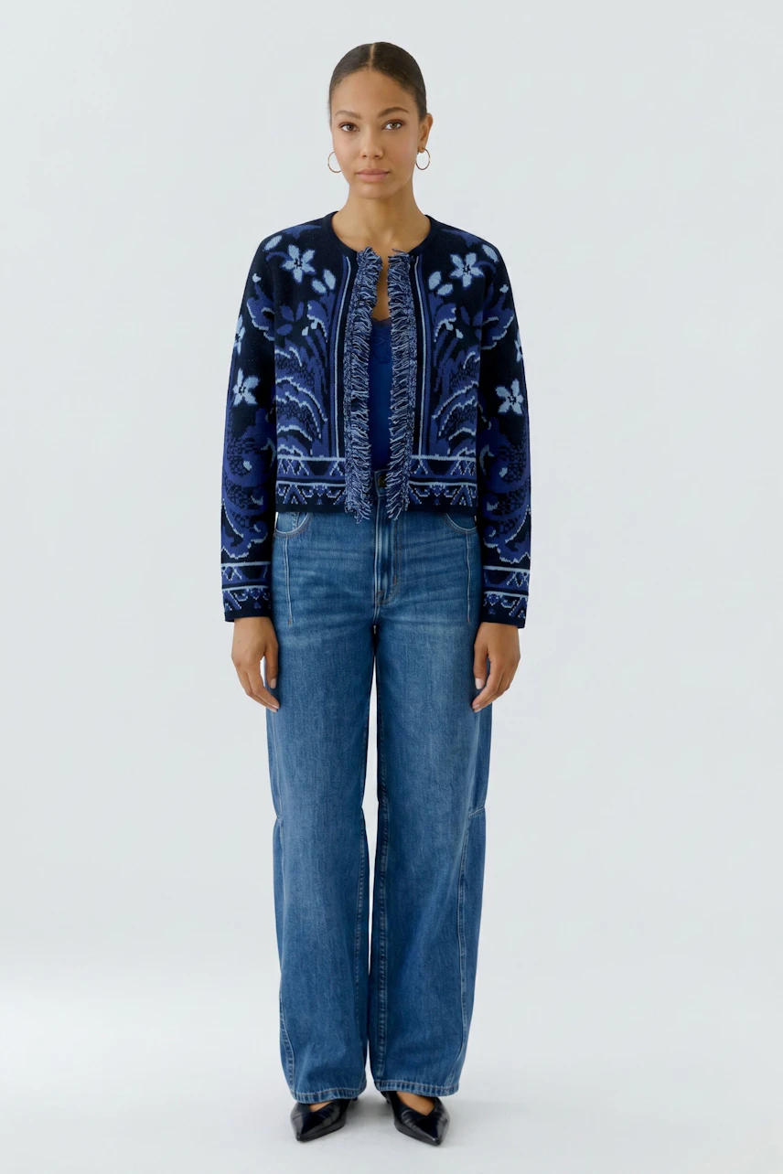Frau in blauer Strickjacke mit floralen Mustern, kombiniert mit Jeans und schwarzen Schuhen.