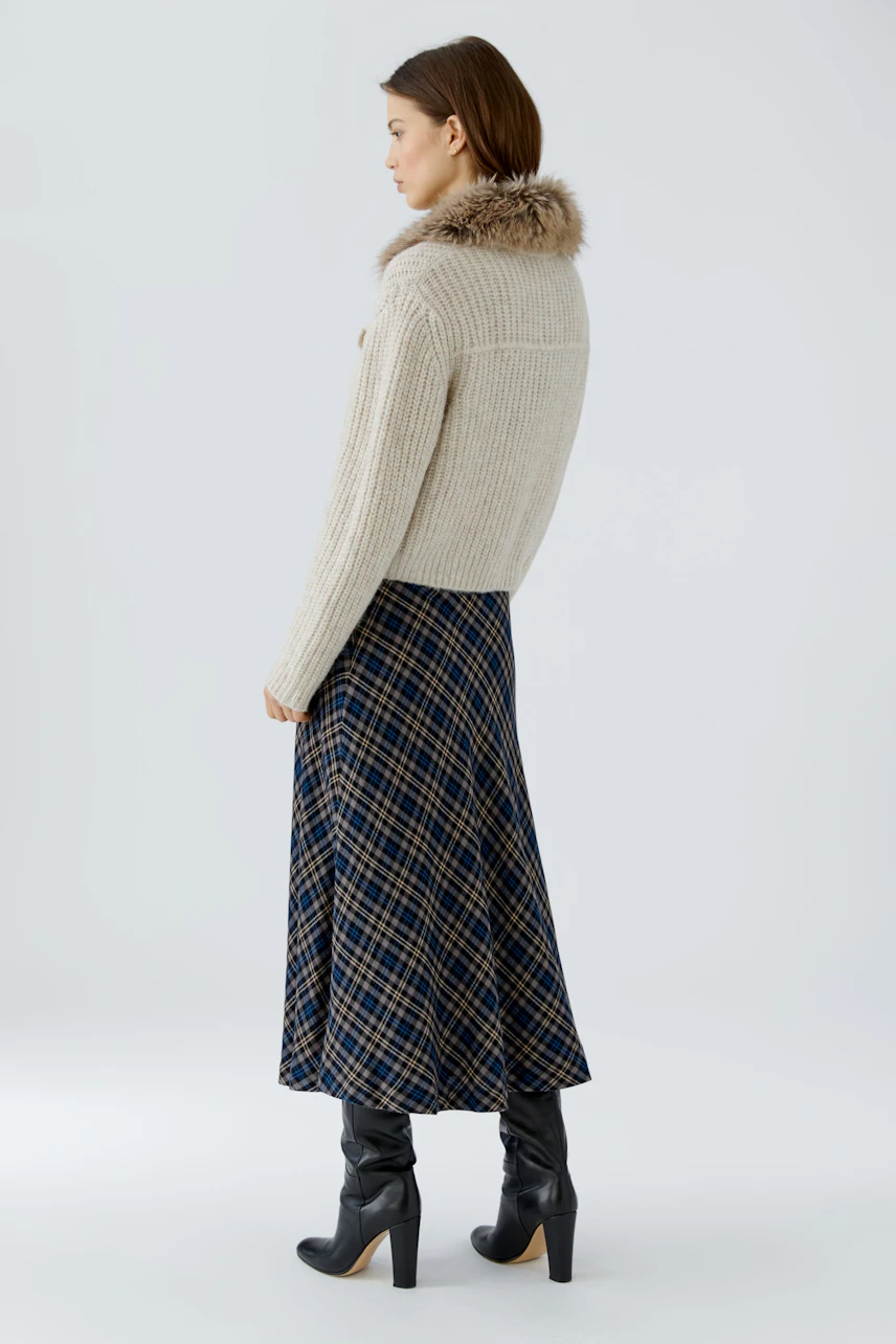 Woman in beige cardigan with fur collar, paired with plaid skirt and black boots, side view.