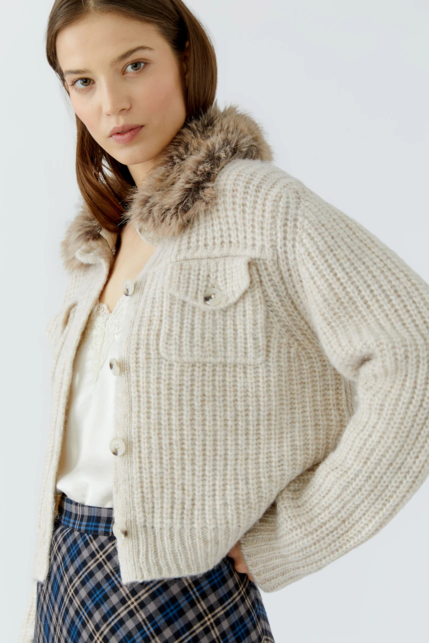 Woman in cozy cardigan: Beige knit cardigan with faux fur collar, paired with a plaid skirt and white top.
