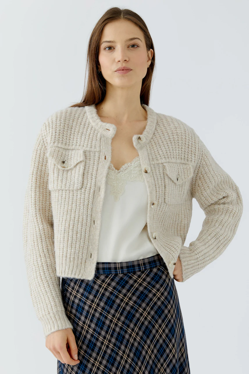 Woman in cream knit cardigan, lace top, and plaid skirt, exuding a casual and confident style.