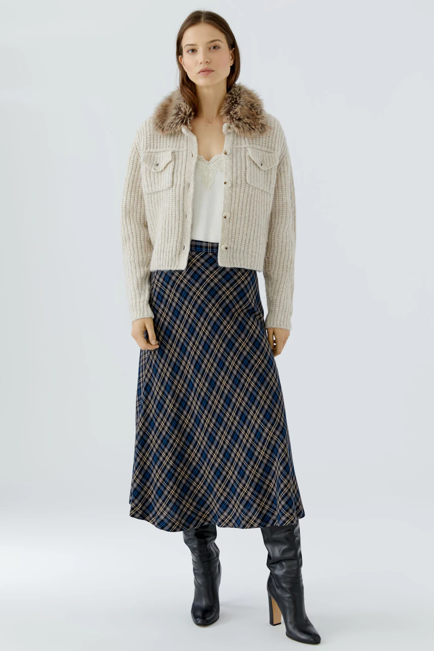 Woman in a cozy cardigan with fur collar, plaid skirt, and boots, exuding a casual style.