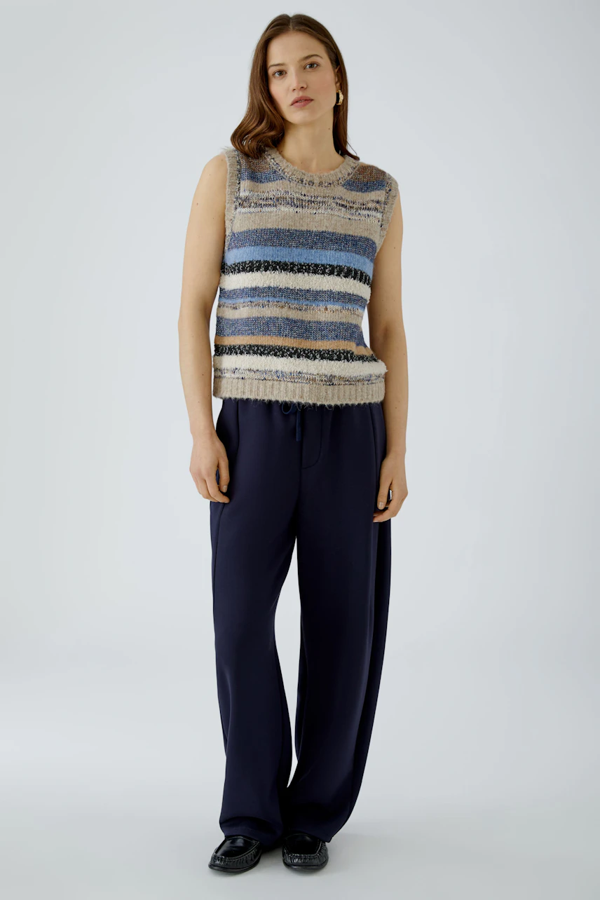 Casual women's pullover: Woman in a striped sleeveless pullover with navy trousers, standing confidently.