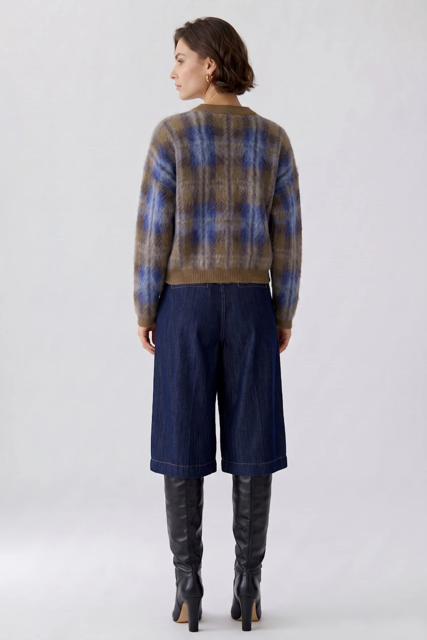 Woman in plaid pullover, blue and brown tones, paired with denim culottes and knee-high boots, facing away.