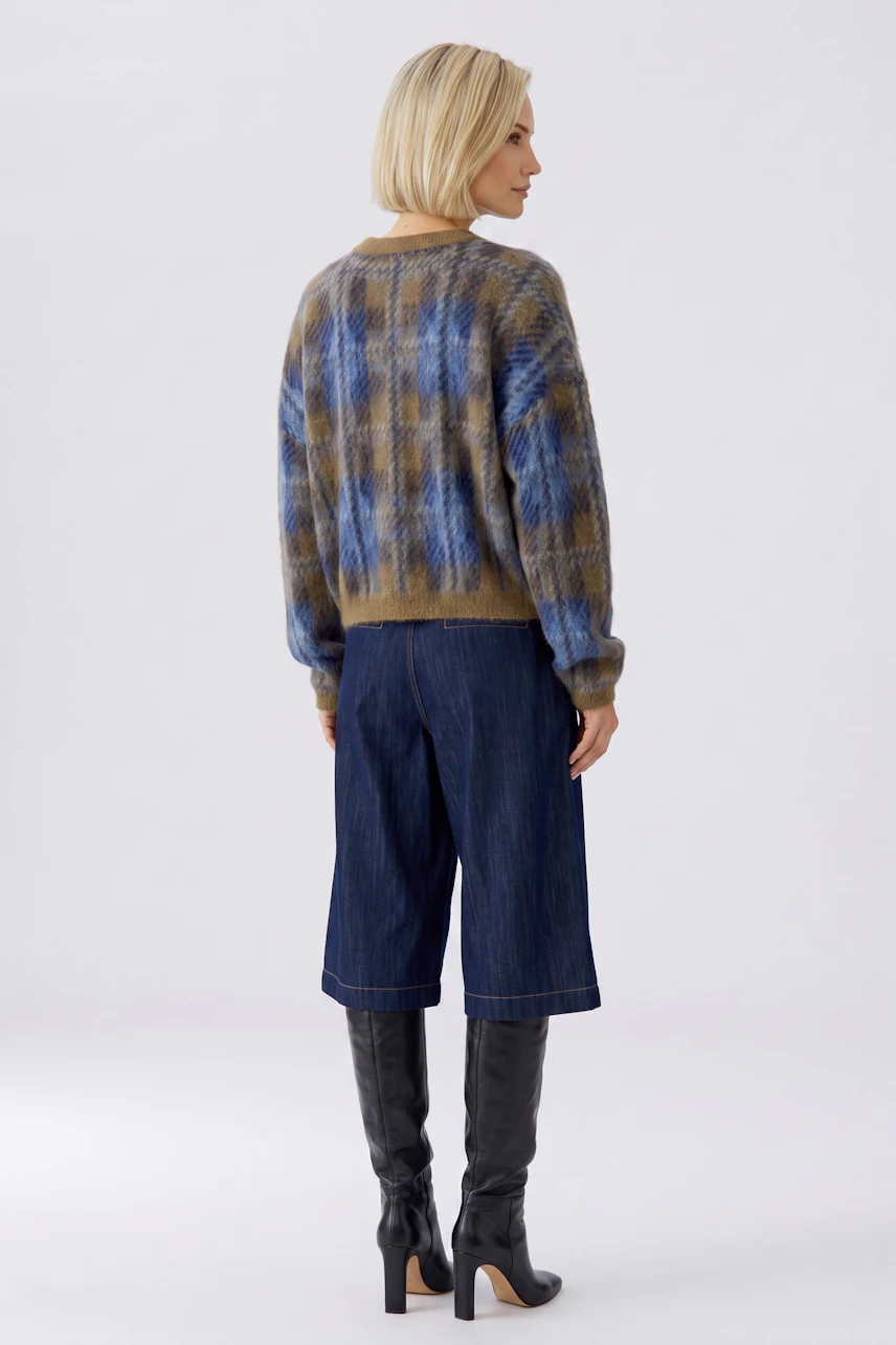 Woman in plaid pullover, blue and brown tones, paired with denim culottes and knee-high boots, facing away.