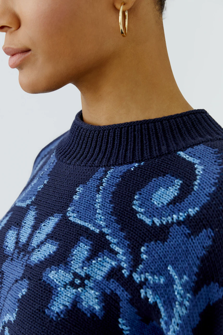 Frau im blauen Pullover mit floralem Muster und goldenen Ohrringen, zeigt stilvolle Details.