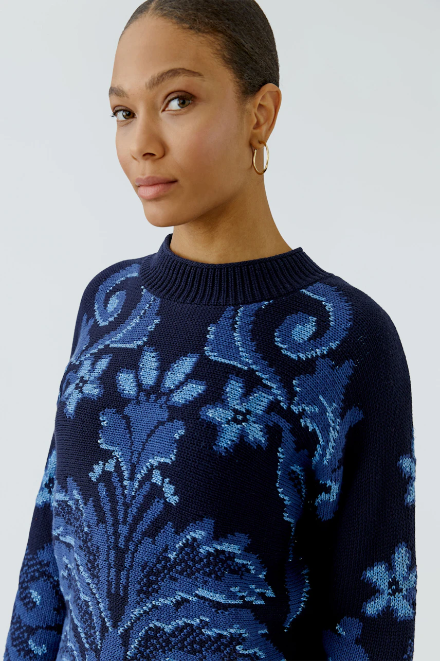 Damen-Pullover: Frau im blauen Pullover mit floralem Muster und Rundhalsausschnitt schaut selbstbewusst.