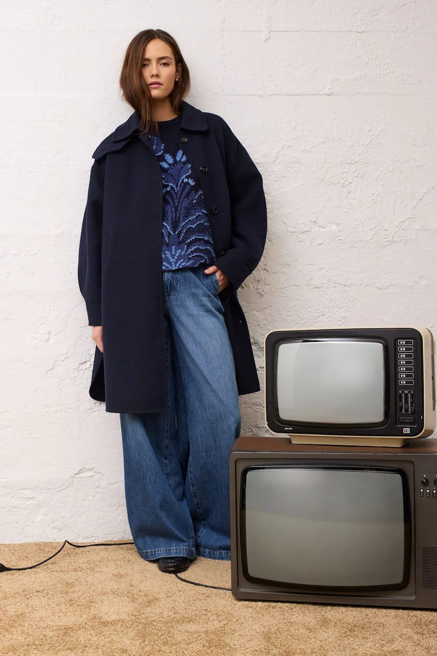 Casual pullover: Woman in a blue patterned pullover, wide jeans, and a dark coat stands by vintage TVs.
