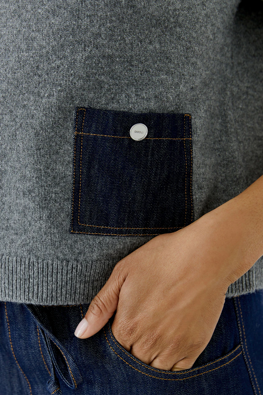 Casual pullover: Woman in a grey pullover with a denim pocket detail, hand resting on hip.