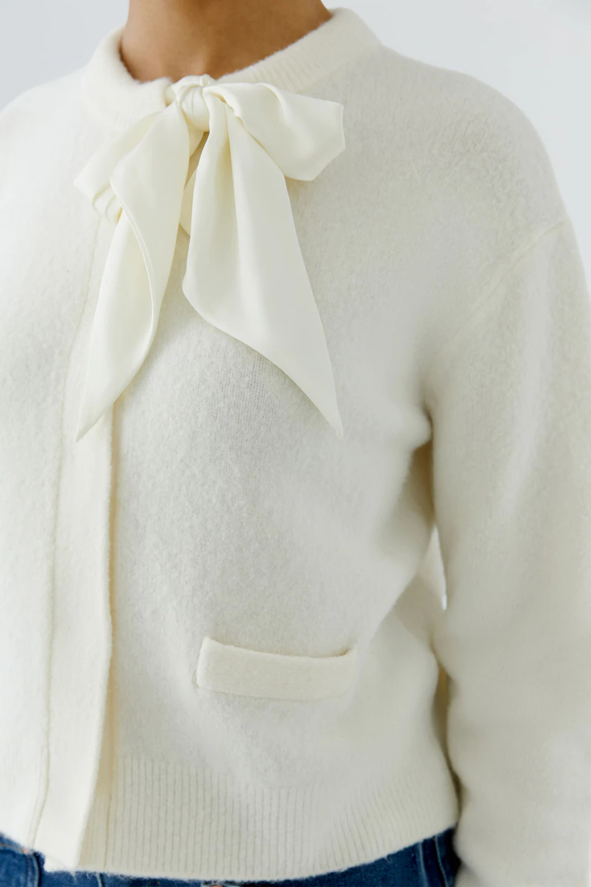 Woman in cream cardigan with bow detail, paired with blue jeans. Casual and stylish look.