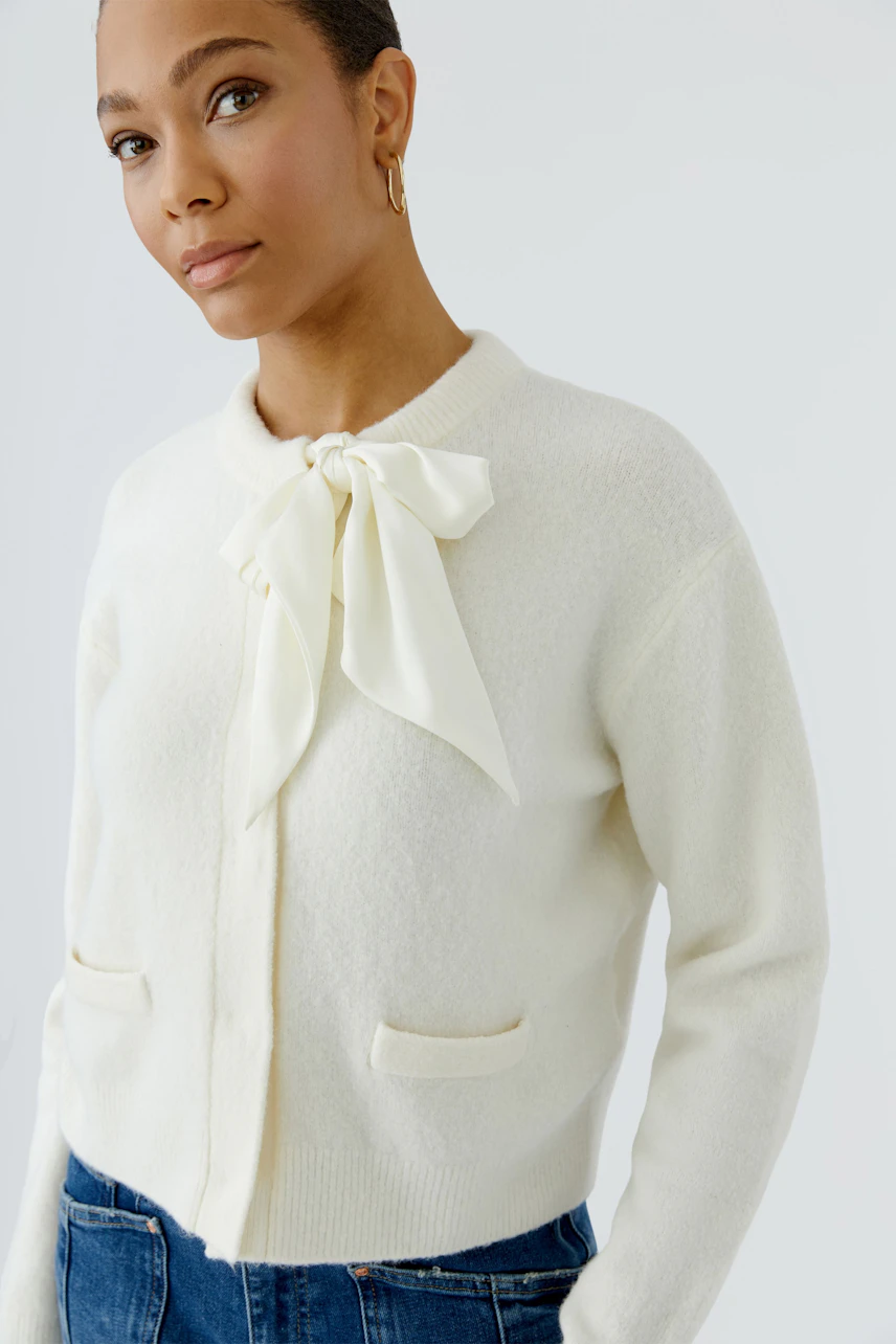 Woman in white cardigan with bow detail, paired with blue jeans, looking confidently.
