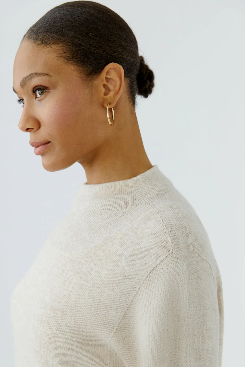 Woman in a beige pullover with a simple design, looking to the side. Pullover has a high neckline.