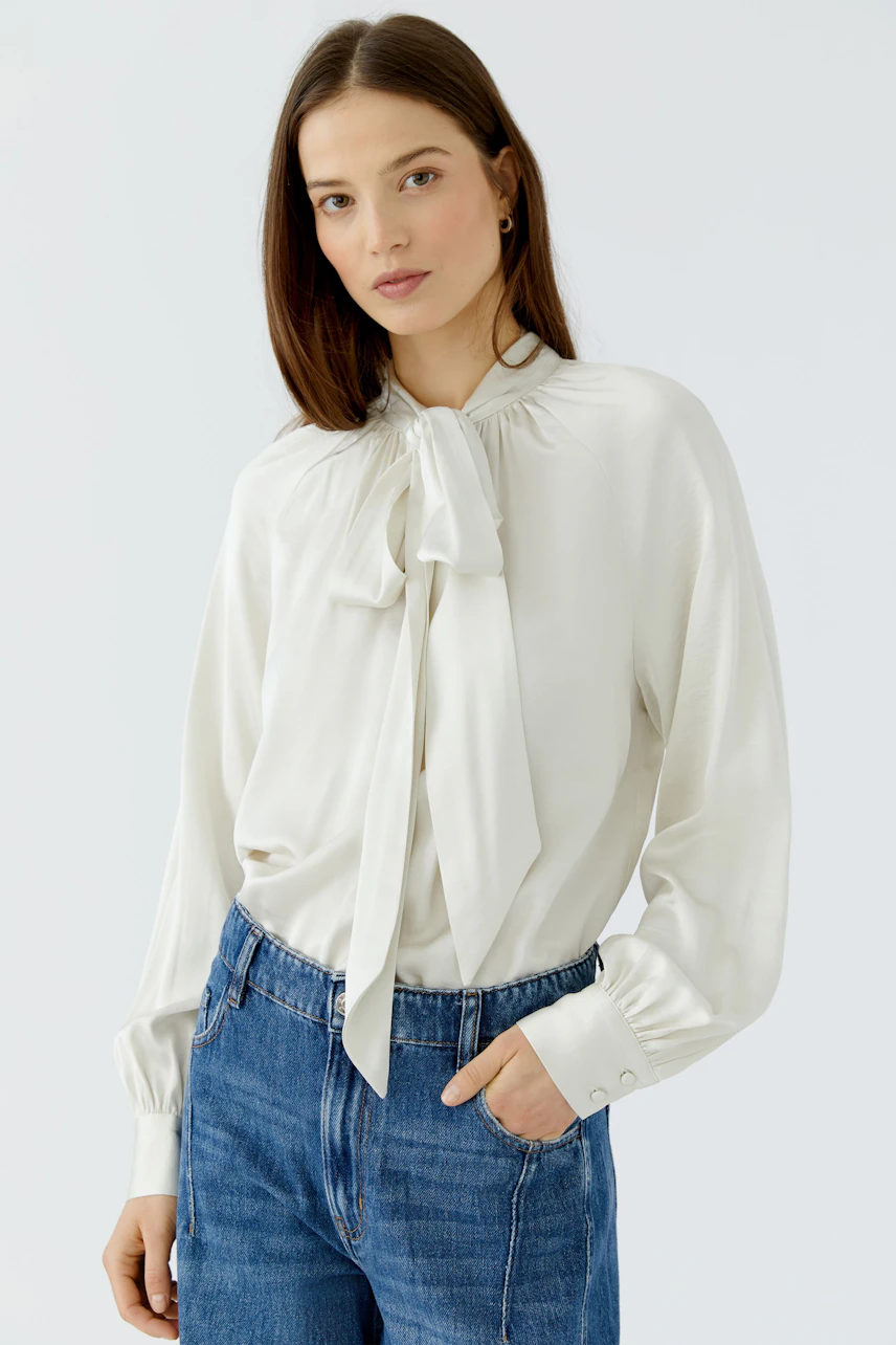 Woman in white pussy-bow blouse and blue jeans, hands in pockets, looking confidently.