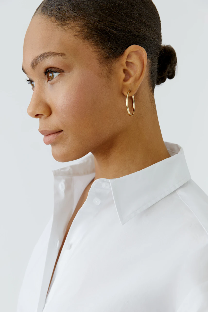 Woman in a crisp white shirt blouse with gold hoop earrings, looking to the side.