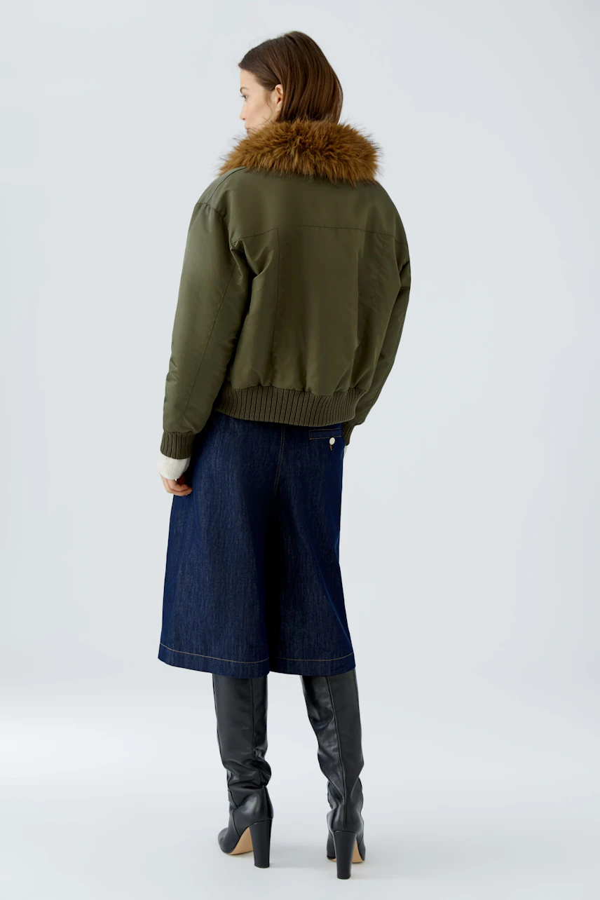 Woman in green blouson with fur collar, denim skirt, and black boots, facing away.