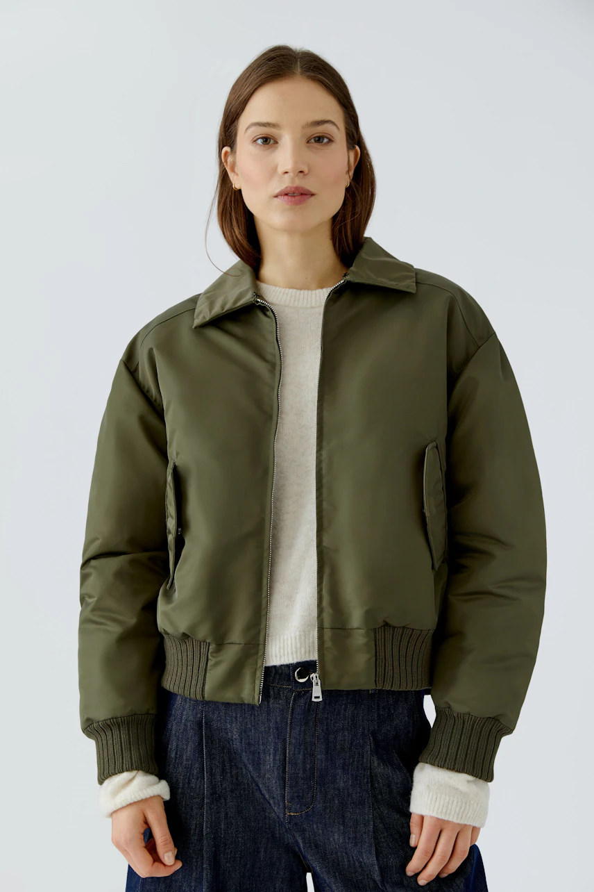 Casual blouson: Woman in olive green blouson, white pullover, and dark jeans, looking confidently.